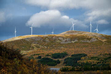 Sortland, scenic landscape featuring wind turbines on a hill with autumn foliage, set against a partly cloudy sky, Norway