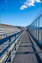 Tromsobrua Bridge long, empty bridge with a metal railing and barbed wire on the right, set against a clear blue sky and distant mountains, Tromso Norway