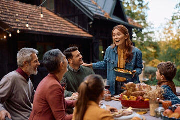 Happy woman brining food at table while celebrating Thanksgiving with her extended family.
