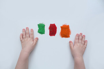 Child's hands next to three piles of colored sand: green, red, orange, on a white background. Child's creativity.