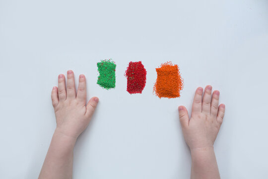 Child's hands next to three piles of colored sand: green, red, orange, on a white background. Child's creativity.