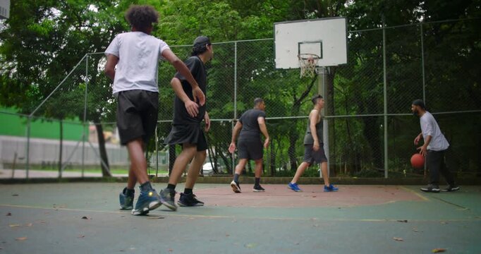 Hispanic man helping friend of African descent up from ground after fall on outdoor basketball court, teammates reuniting to continue friendly street game