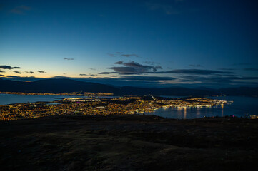 A breathtaking night view of Tromso, Norway nestled between a mountainous landscape and a calm body of water, with city lights reflecting on the water, at the top of Storsteinen