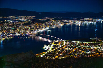 A stunning night view of Tromso, Norway, illuminated by lights, with a harbor and a bridge stretching across the water, at the top of Storsteinen