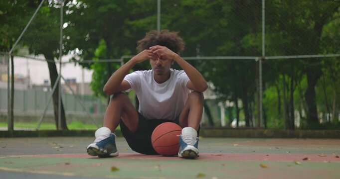 Upset African American young man sitting on basketball court holding basketball between legs, head down in frustration, expressing defeat and inner conflict after game