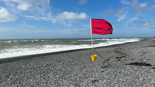 Red flag warning flag blowing in strong wind on stone beach with rough waves