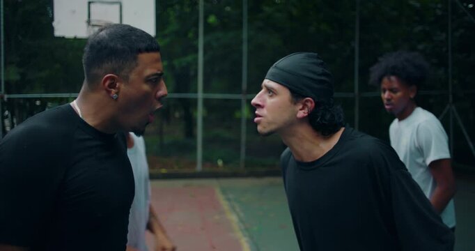 Young men of African and Latin descent in heated argument on outdoor basketball court, yelling and pushing each other face to face in tense moment of anger and disagreement