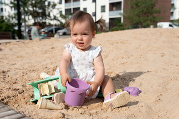 delighted toddler with pigtails joyfully playing in a sunlit urban sandbox, surrounded by colorful toys, with modern apartment buildings and blurred greenery in the background, dynamic, joyful
