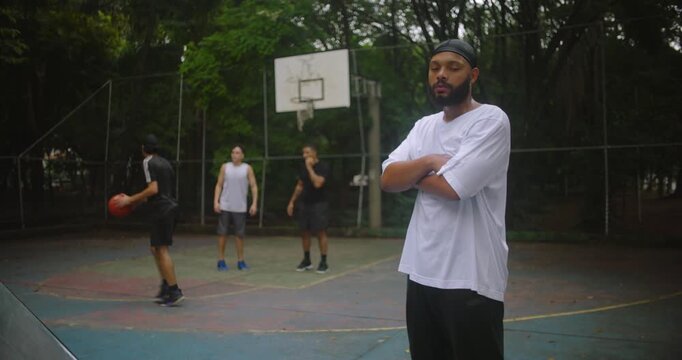 African descent man wearing durag with arms crossed on basketball court looking confident and focused while friends play behind, symbolizing leadership, resilience, and strength in teamwork