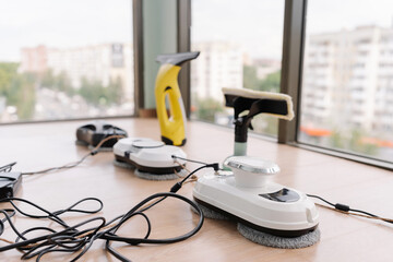 Several window cleaning robots, cordless window cleaner and window cleaning mop sitting on ledge, cables attached and ready for cleaning large window with view of city, close-up, no people.