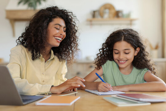 Young mother helping daughter with school homework while working on laptop, sitting together at kitchen table. Girl writing in notebook, learn with freelance woman