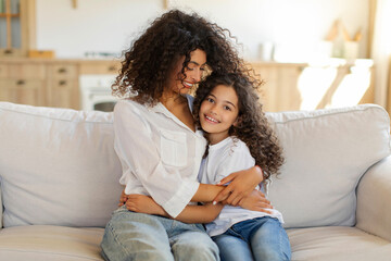 Loving Latin family mother and daughter bonding at home, woman and girl embracing, sitting on couch and smiling, copy space