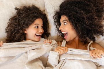 Funny mother and her preteen daughter lying under blanket, having fun and spending morning in bed, looking at each other and smiling, top view