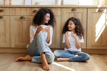 Young mother and preteen daughter drinking tea and chatting while sitting on floor at kitchen, full...