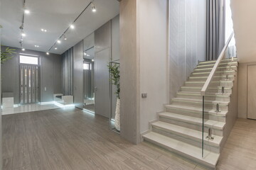 A stylish hallway showcases a sleek staircase and modern decor. Natural light filters through large windows, highlighting clean lines and minimalist furnishings in the setting.