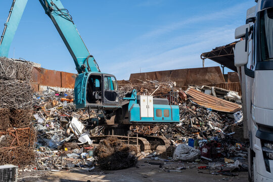 Excavator loading scrap metal onto truck in junkyard. Industrial excavator working at metal recycling yard. Excavator handling metal waste at recycling facility.