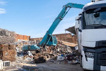 Excavator loading scrap metal onto truck in junkyard. Industrial excavator working at metal recycling yard. Excavator handling metal waste at recycling facility.