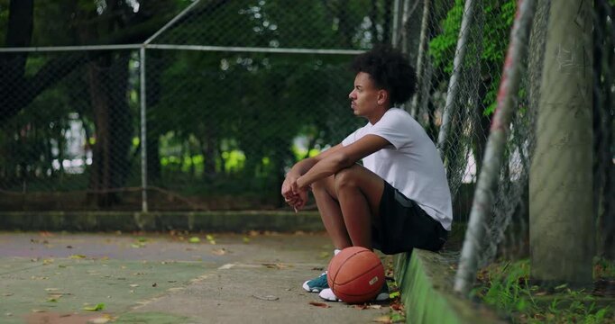 African American young man sitting by fence on outdoor basketball court, resting with ball at feet, expression of contemplation, focus, and perseverance after training