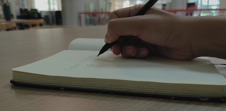 A person's hands, actively writing notes with a black pen on a blank page of a small, open notebook on a wooden table. 