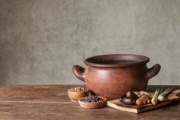 An antique clay cauldron and spices on a wooden table with a concrete wall in the background. A rustic, vintage-style background.