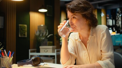 Dedicated manager in a corporation stays after hours to focus on budgeting, organizing and scheduling the next phase of project development. Woman drinking a glass of water at desk. Camera A.