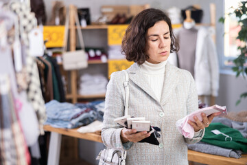 Adult woman in a blazer holds sets of cotton socks in hands and chooses between them, she stands in a clothing store against the background of a shelf and a counter