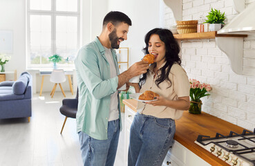 Romantic breakfast of happy couple in home cozy kitchen, smiling man sharing morning croissant, feeding woman. Young cheerful people holding plates and coffee mugs, enjoying with smiles time together