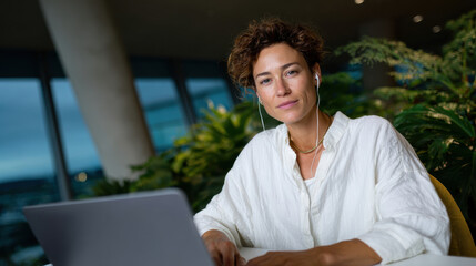 A focused businesswoman with headphones works gracefully in a modern office setup, embodying the essence of productivity and creativity in a serene, plant-filled environment.