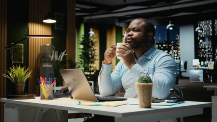 Black office employee serving a cup of coffee and doing overtime at workplace, staying awake for budgeting tasks in the strategy room after hours. Caffeine refreshment for proofreading.