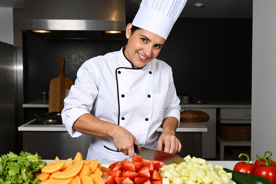 Happy female chef in a white uniform and hat smiling while chopping fresh vegetables in a professional restaurant kitchen. Culinary professional preparing ingredients for a gourmet meal - Powered by Adobe