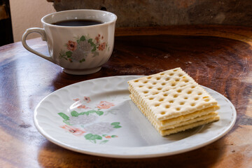 Floral ceramic cup of coffee and plate of crackers on rustic wooden surface, capturing cozy morning snack setup with elegant dishware and warm atmosphere.