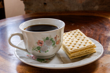 Cup of black coffee with floral design and stack of crackers on saucer, placed on rustic wooden table in cozy morning setup with warm, homely atmosphere.