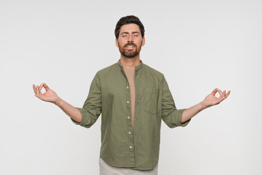 Young tranquil man in casual clothes standing isolated on white background makes mudra gesture meditates feels internal balance having healthy habit, wellbeing harmony no stress concept
