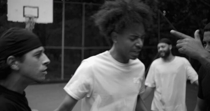 Heated argument between two men of Latin and African descent on outdoor basketball court, face to face in conflict as teammate steps in to defuse tension, black and white image