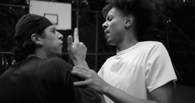 Hispanic and African descent men in heated argument on outdoor basketball court, shoving each other as tempers rise and emotions escalate, black and white
