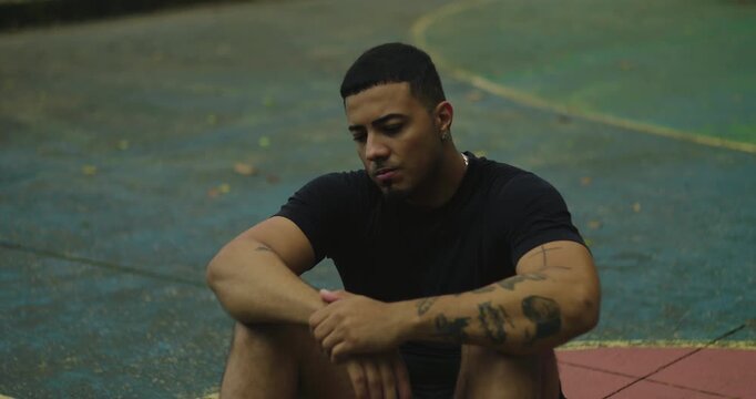 Latin Hispanic man of African descent sitting on empty basketball court, hand behind head, looking contemplative and concerned, showing introspection and emotional exhaustion after exercise