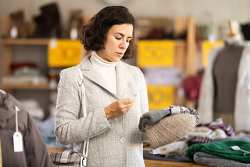 Adult woman stands in a store against the background of a counter with clothes and chooses a...