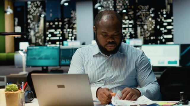 Confident male employee working on a new project task after hours, sitting alone in dark office late at night. Guy fact checking information and monthly agenda for objectives. Camera B.