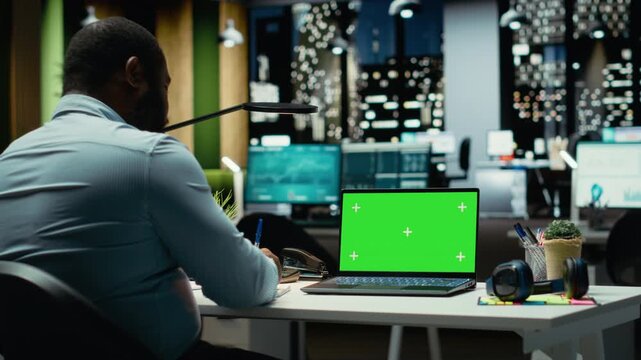 Black male worker looks at chroma key screen on laptop at night, solving business tasks after hours. Office consultant working on project development, meeting targets and goals. Camera B.