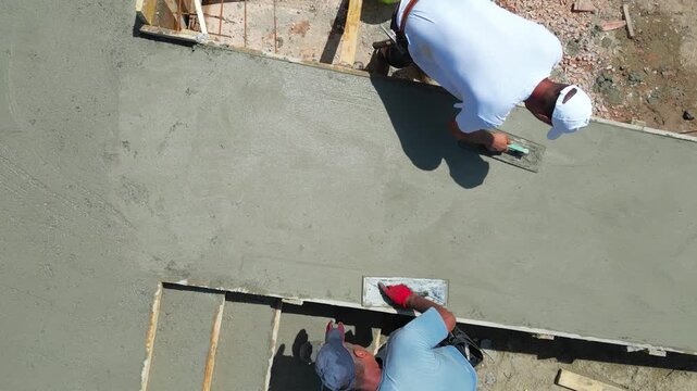 Construction workers are using finishing trowels on freshly poured concrete to create a smooth, level surface at a building site. View from above shows workers leveling the surface of the stairs