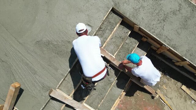 Construction workers are using finishing trowels on freshly poured concrete to create a smooth, level surface at a building site. View from above shows workers leveling the surface of the stairs
