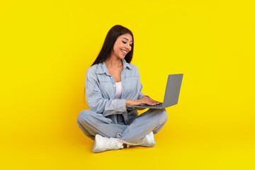 Naklejka premium A woman sits comfortably on the floor, legs crossed, with a joyful expression while typing on her laptop. The bright yellow backdrop adds a cheerful vibe to the scene.