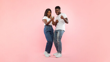A black couple stands in front of a pink background, happily celebrating their success. They share a smartphone, shaking their fists in excitement.