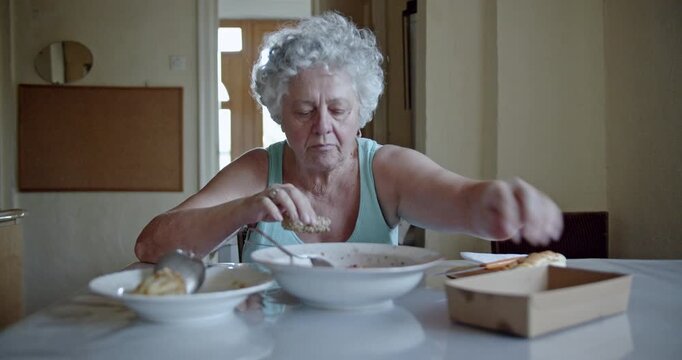 A mature senior woman with curly grey hair carefully breaks a piece of bread before thoughtfully eating her meal from a bowl, highlighting a moment of daily life in her home.