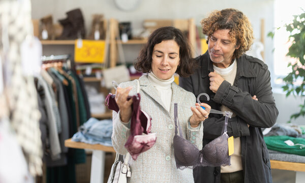 Adult woman chooses a comfortable bra, next to a man who gives recommendations. Husband and wife buy underwear from a clothing store