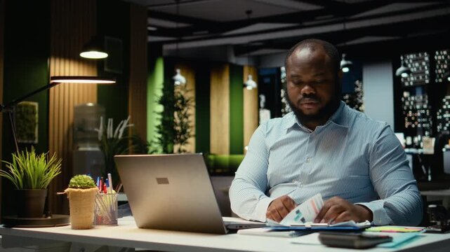 Male company analyst reading data reports and fact checking on laptop, planning project and scheduling tasks after hours. Notetaking and handwriting for objectives achievement. Camera A.