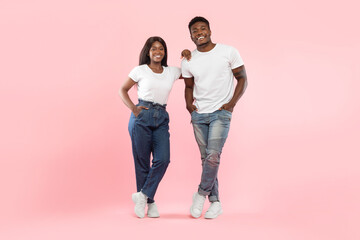 Young African American couple stands confidently in a pink studio. The lady leans on the guy, both smiling and looking at the camera. They wear casual outfits and display happy energy.