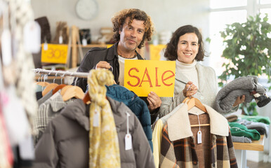 Happy couple, woman and man holding sign saying Sale for winter season standing in interior of modern clothing boutique