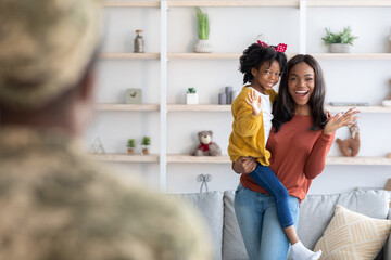 A mother holds her young daughter while joyfully greeting a soldier returning home. The cozy living room features plants and decorations, creating a warm atmosphere.