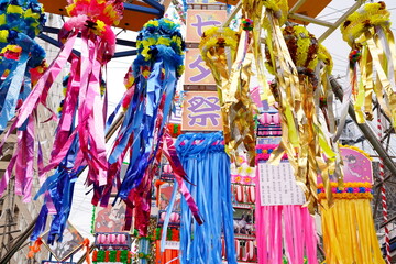 色とりどりの紙テープや布でできた七夕飾りが頭上を覆い尽くすように吊り下げられている賑やかな商店街の様子です。ピンク青金色など派手な装飾が印象的です。